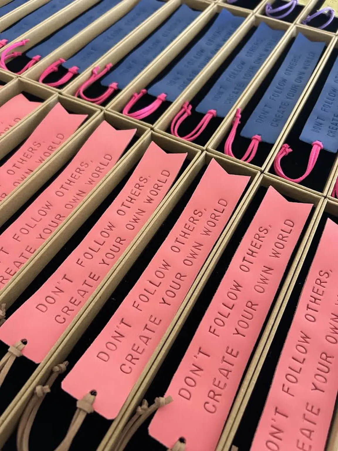 Marcadores de livros em couro rosa e azul com frases gravadas, organizados em caixas individuais.