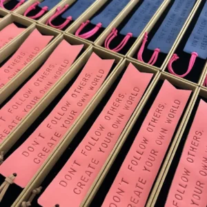 Marcadores de livros em couro rosa e azul com frases gravadas, organizados em caixas individuais.