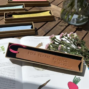 Marcador de livros em couro castanho com frase gravada, dentro de caixa, sobre livro aberto com flores ao lado.