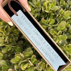 Marcador de livros em couro azul-claro com frase gravada, dentro de caixa, segurado à frente de plantas verdes.