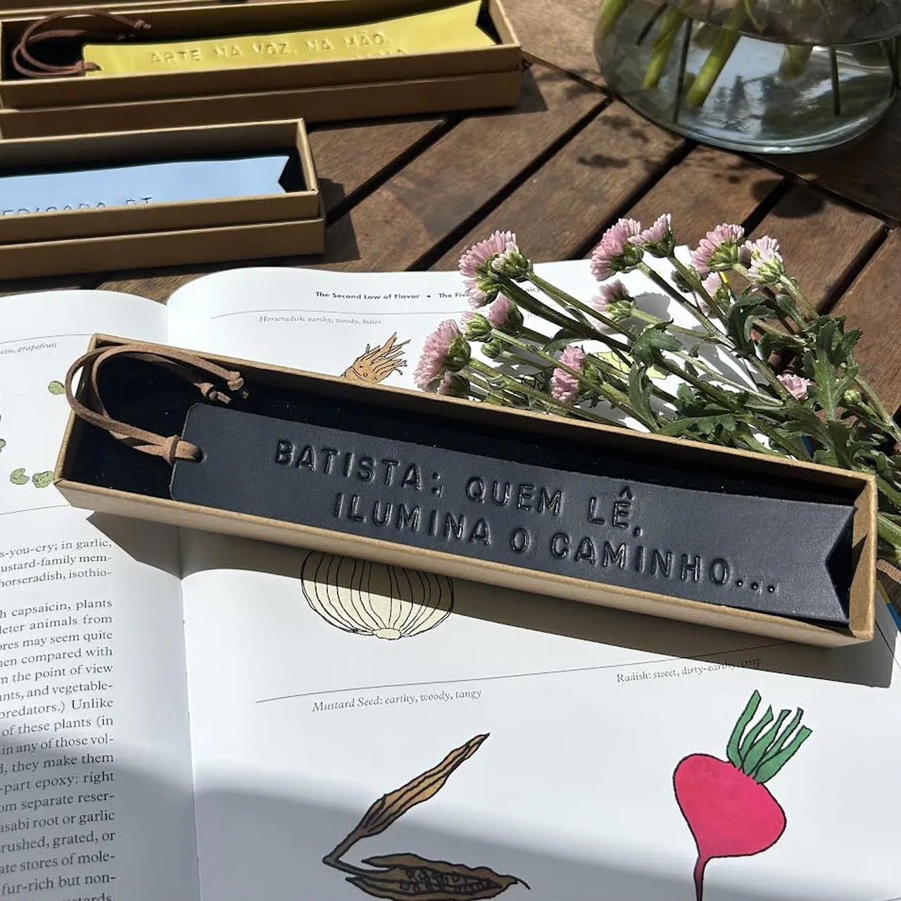 Marcador de livros em couro preto com frase gravada, apresentado numa caixa sobre um livro aberto com flores ao lado.