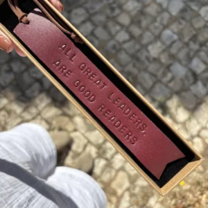 Marcador de livros em couro bordô com frase gravada, apresentado numa caixa sobre calçada portuguesa.