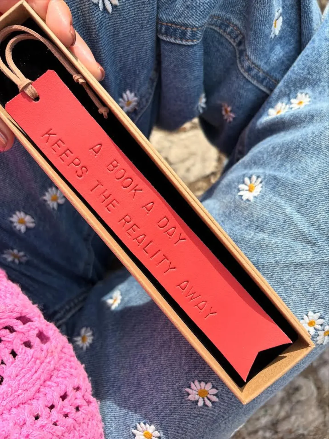 Marcador de livros em couro vermelho com frase gravada, apresentado numa caixa sobre roupa de ganga com bordados florais.