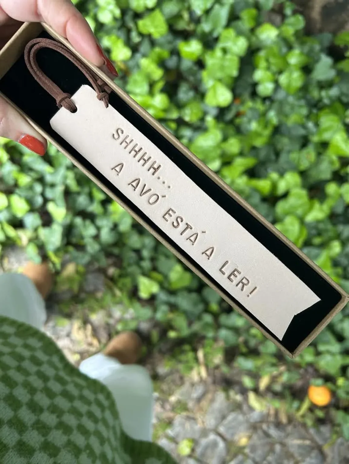 Marcador de livros em couro claro com frase gravada, exibido numa caixa sobre fundo de folhas verdes.