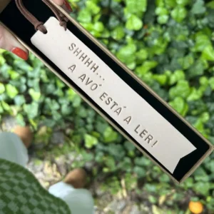Marcador de livros em couro claro com frase gravada, exibido numa caixa sobre fundo de folhas verdes.