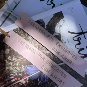 Dois marcadores de livros em couro rosa com frases de agradecimento, colocados sobre revistas e livros.