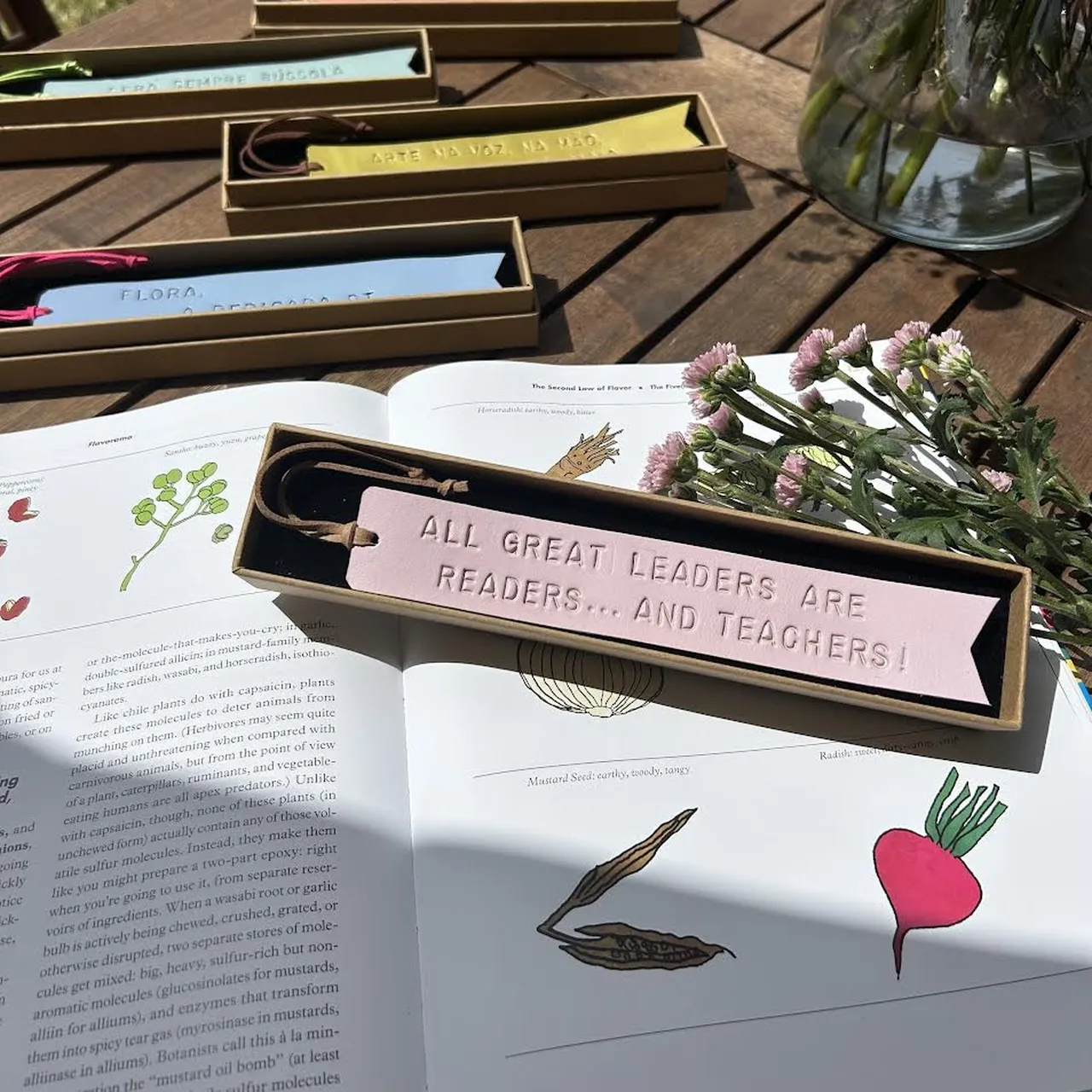 Marcador de livros em couro rosa com frase gravada, colocado numa caixa sobre um livro aberto com flores ao lado.