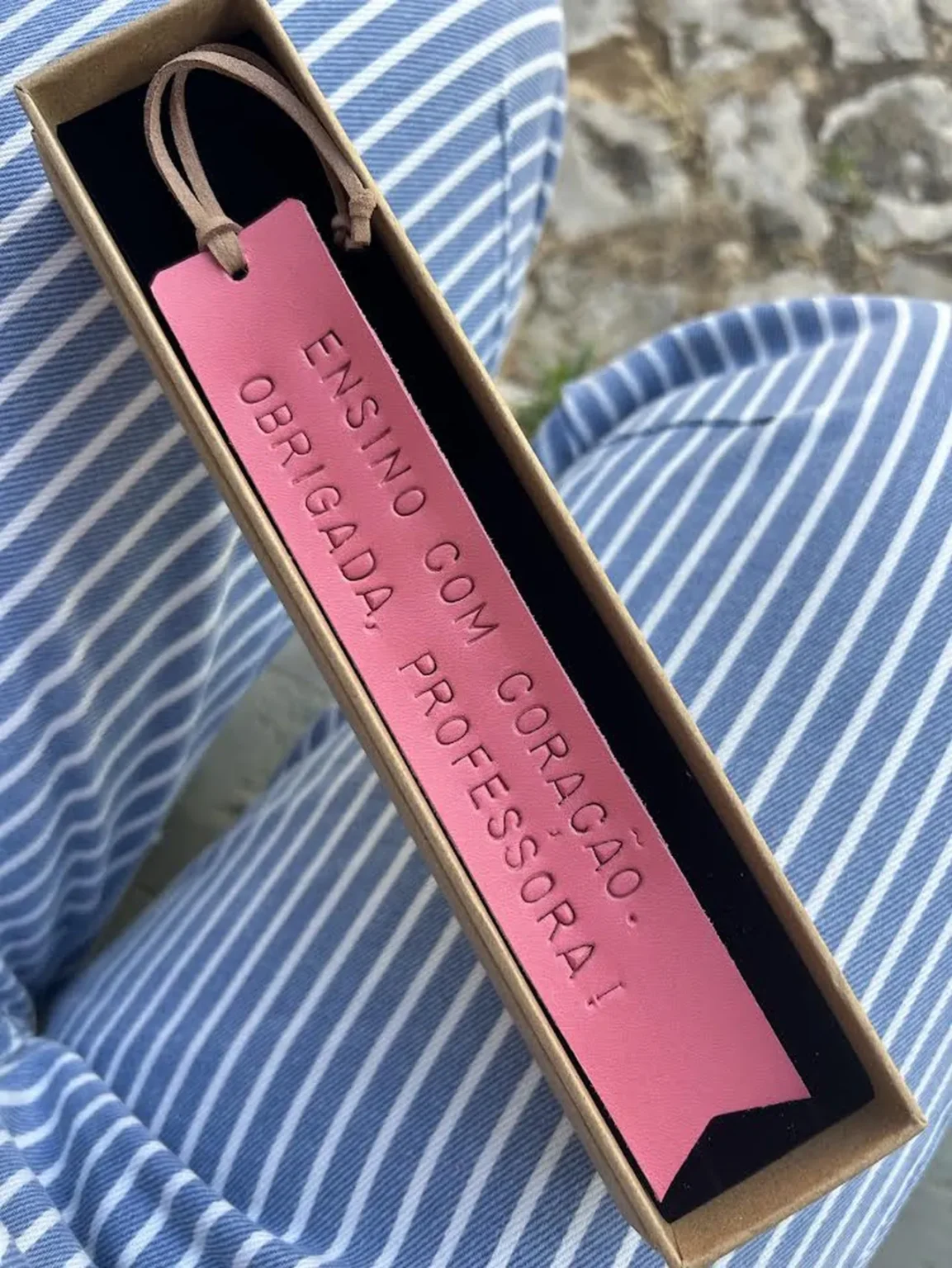 Marcador de livros em couro rosa com frase gravada, apresentado numa caixa sobre tecido às riscas.