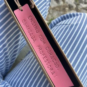 Marcador de livros em couro rosa com frase gravada, apresentado numa caixa sobre tecido às riscas.