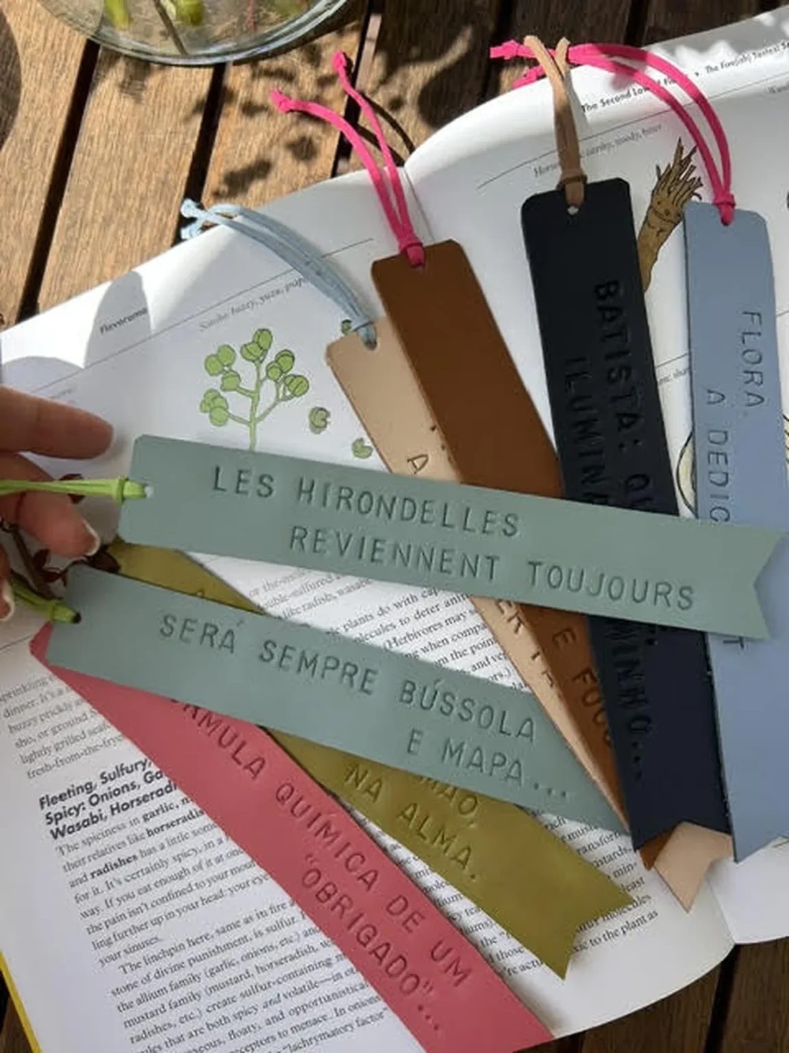 Vários marcadores de livros em couro colorido com frases gravadas, dispostos sobre um livro aberto numa mesa de madeira.