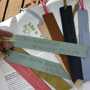 Vários marcadores de livros em couro colorido com frases gravadas, dispostos sobre um livro aberto numa mesa de madeira.