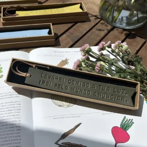 Marcador de livros em couro verde com frase gravada, apresentado em caixa individual sobre um livro aberto com flores ao lado.