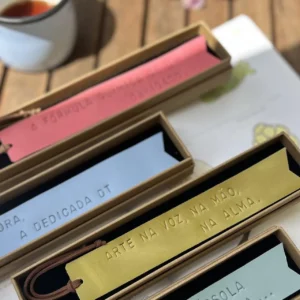 Marcadores de livros em couro colorido com frases gravadas, apresentados em caixas individuais sobre uma mesa de madeira.