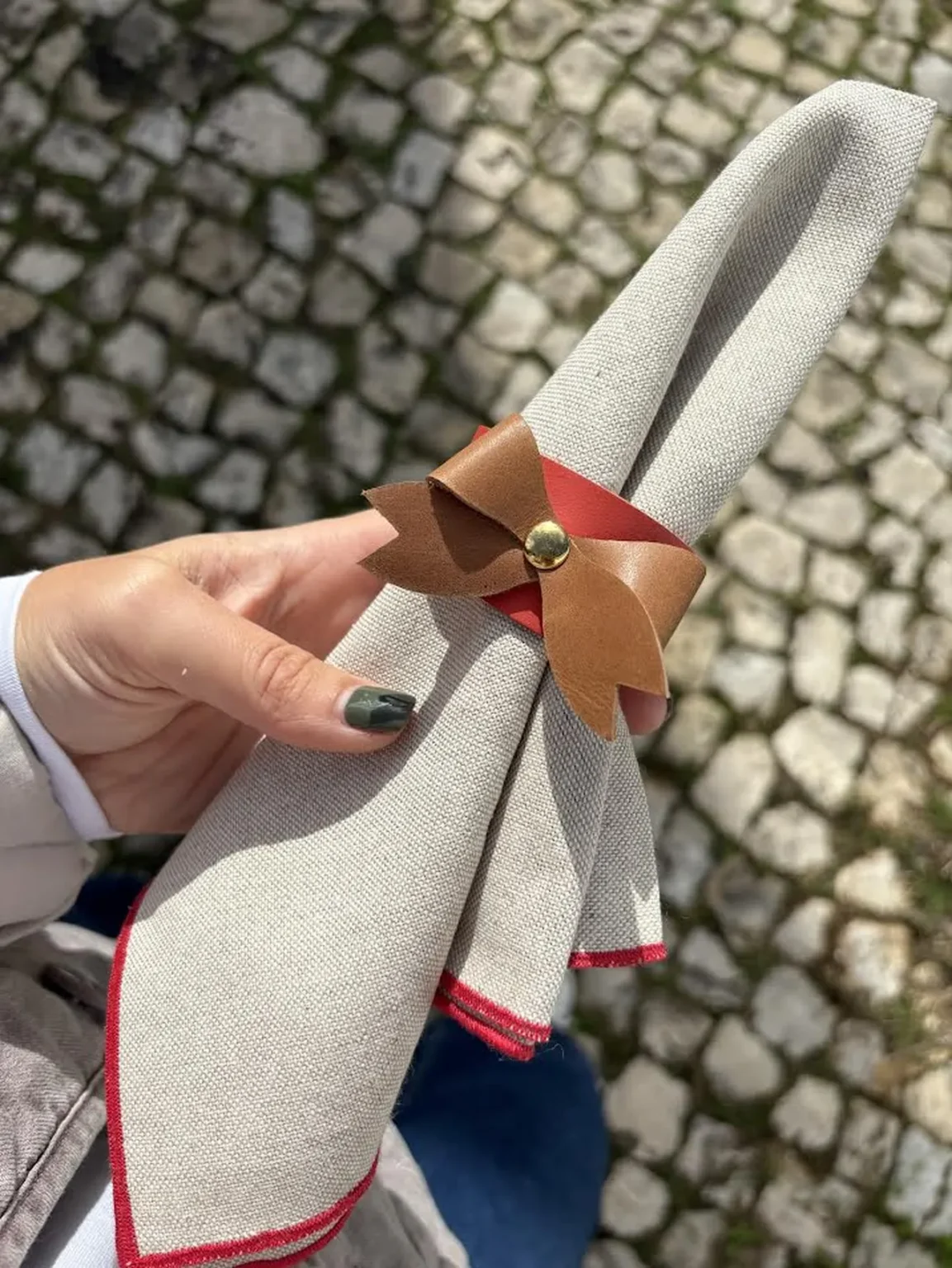 Argola de guardanapo em couro com laço castanho e detalhe vermelho, segurando guardanapo bege com acabamento vermelho.