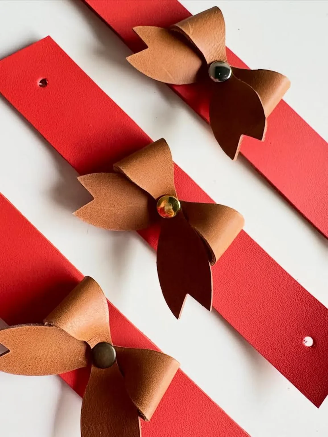 Argolas de guardanapo em couro com laços castanhos sobre tiras vermelhas, com diferentes acabamentos metálicos.