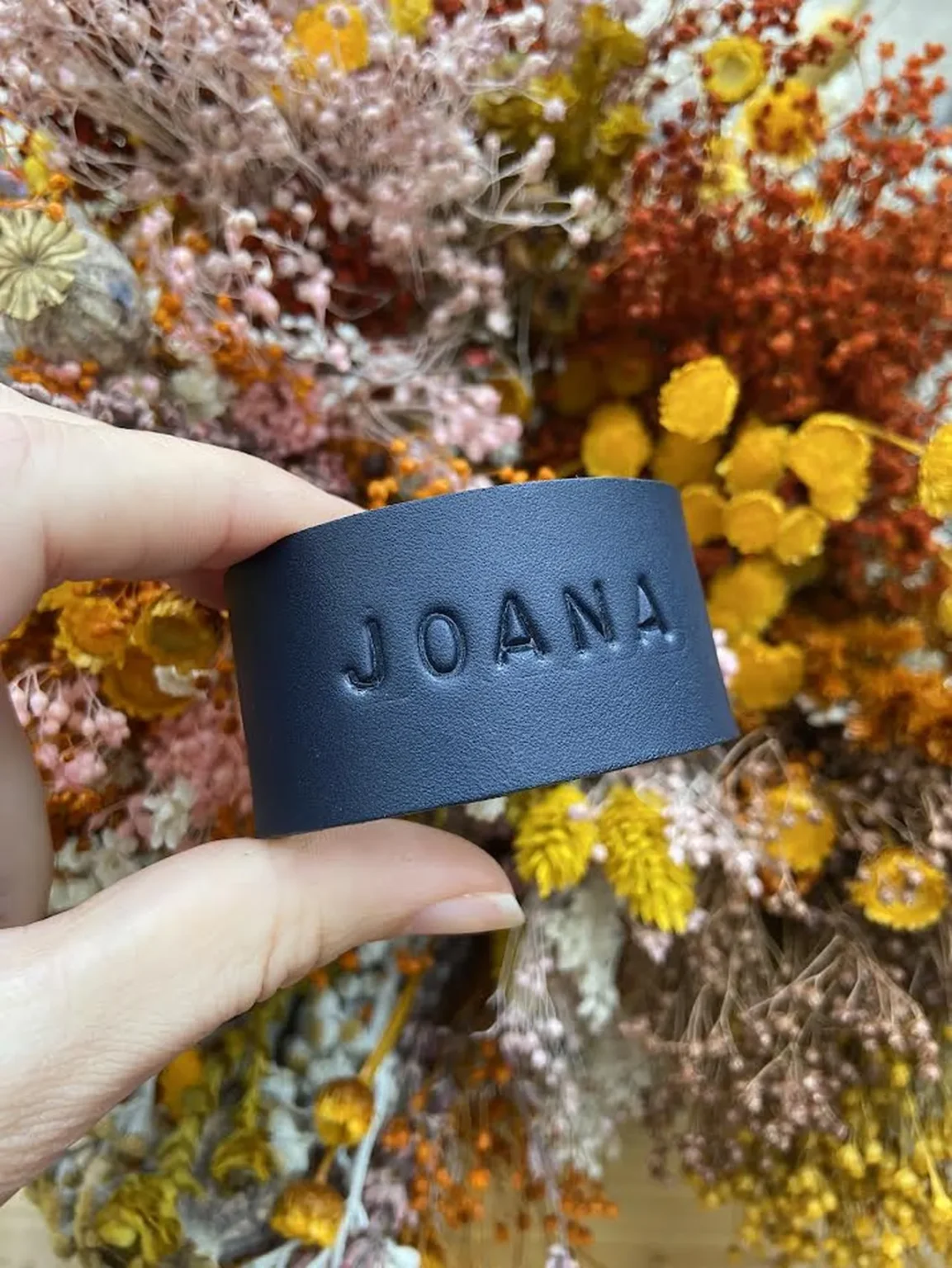 Argola de guardanapo em couro preto personalizada com o nome Joana, segurada à frente de arranjo floral seco.