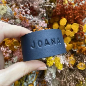 Argola de guardanapo em couro preto personalizada com o nome Joana, segurada à frente de arranjo floral seco.
