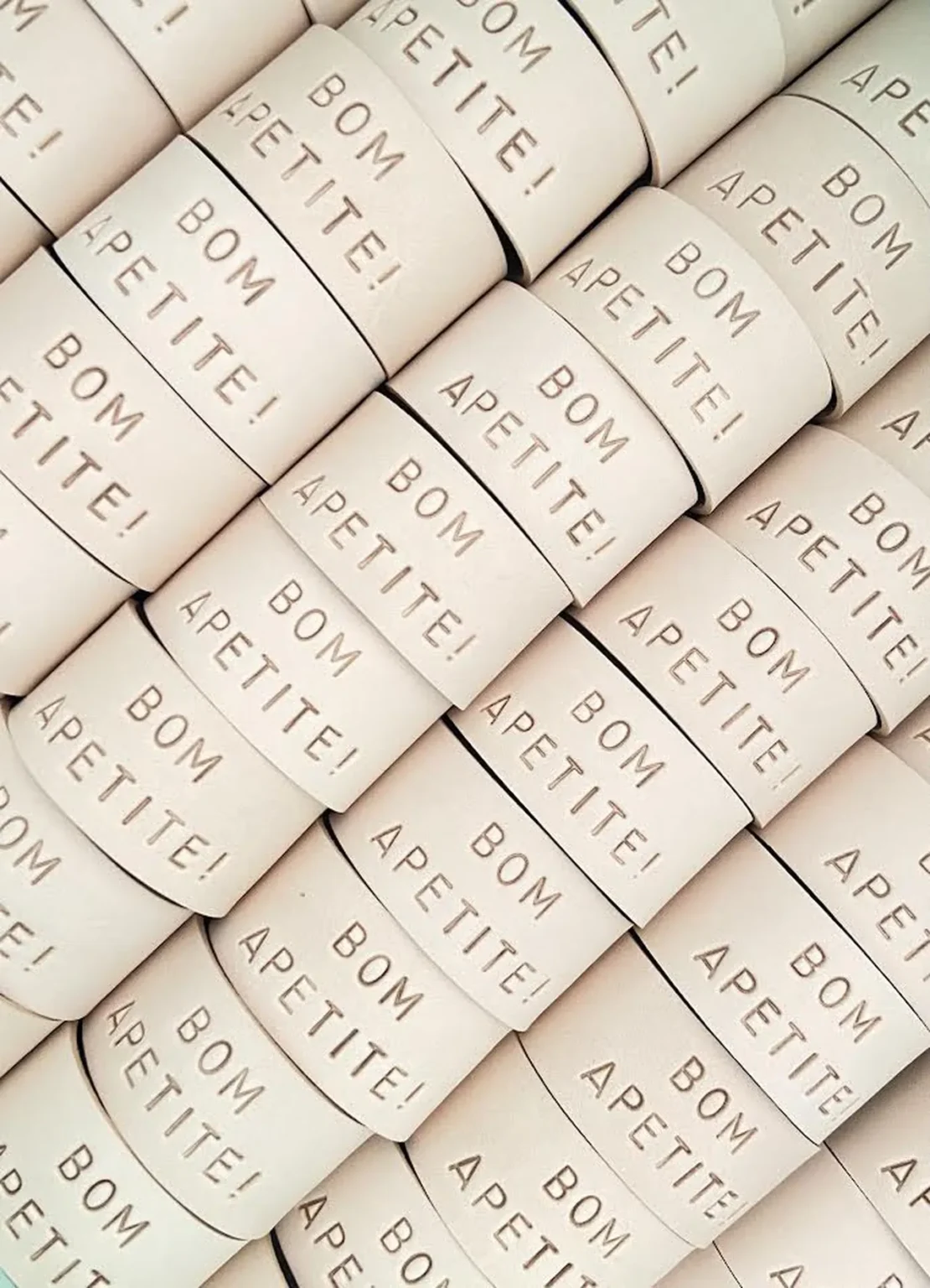 Argolas de guardanapos em couro claro com a gravação “BOM APETITE!”, organizadas em padrão repetido.