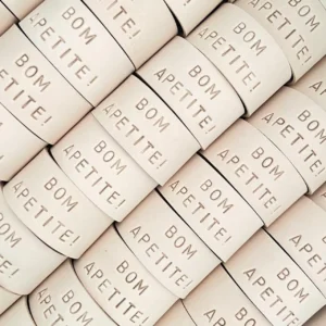 Argolas de guardanapos em couro claro com a gravação “BOM APETITE!”, organizadas em padrão repetido.