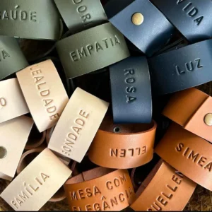 Argolas de guardanapo em couro, em várias cores, com gravações personalizadas de nomes e palavras como “Empatia”, “Luz”, “Bondade” e “Família”, dispostas juntas em destaque.