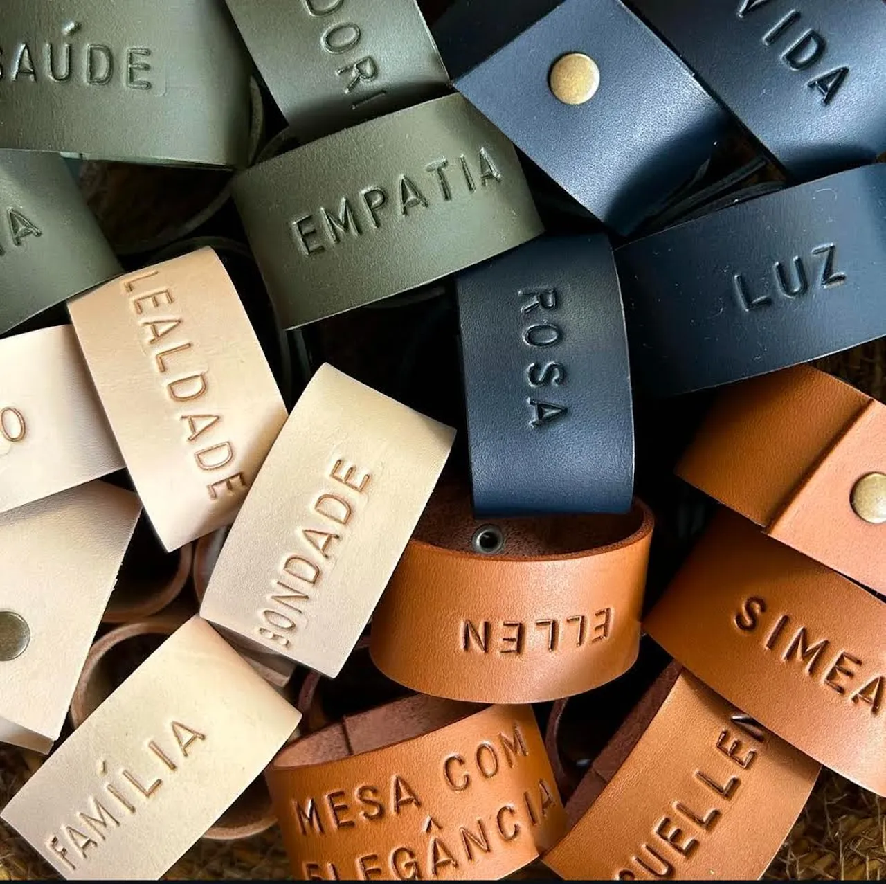 Argolas de guardanapo em couro, em várias cores, com gravações personalizadas de nomes e palavras como “Empatia”, “Luz”, “Bondade” e “Família”, dispostas juntas em destaque.