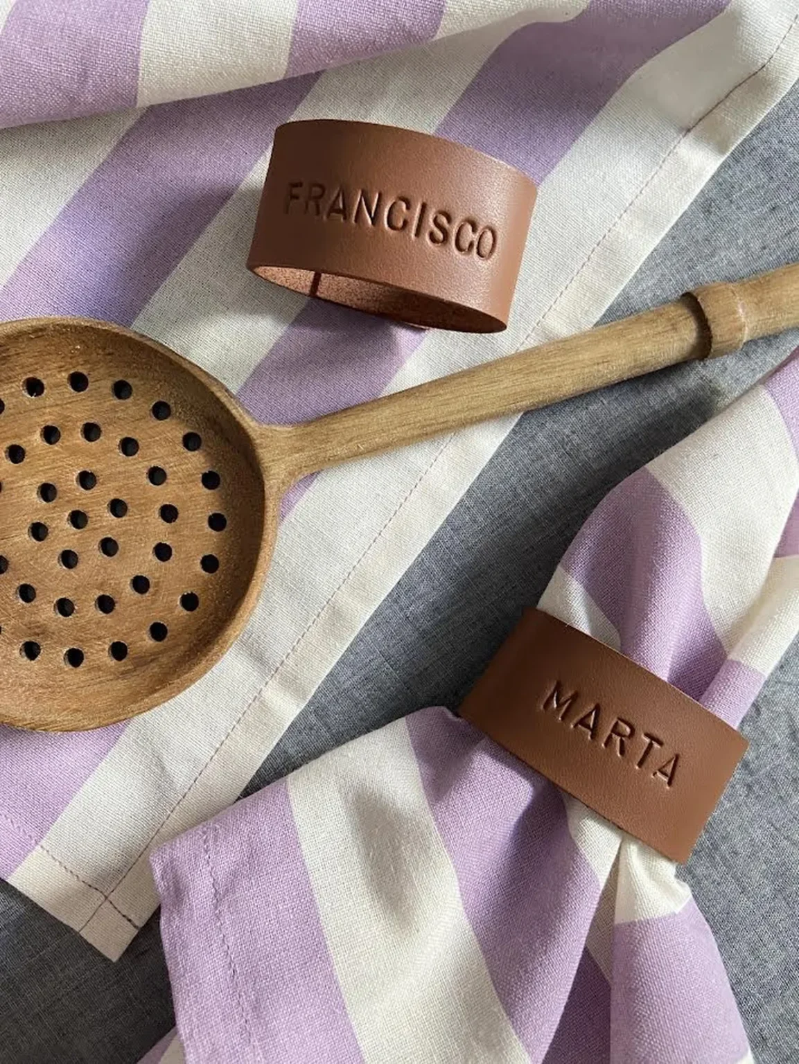 Porta-guardanapos personalizados em pele com os nomes “Francisco” e “Marta”, apresentados sobre guardanapos às riscas lilás e brancas.