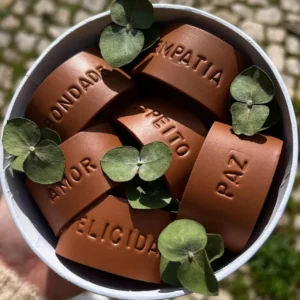 Argolas de guardanapo em couro castanho com palavras gravadas como “Bondade”, “Empatia”, “Amor”, “Respeito”, “Felicidade” e “Paz”, decoradas com pequenas folhas verdes dentro de uma caixa branca.