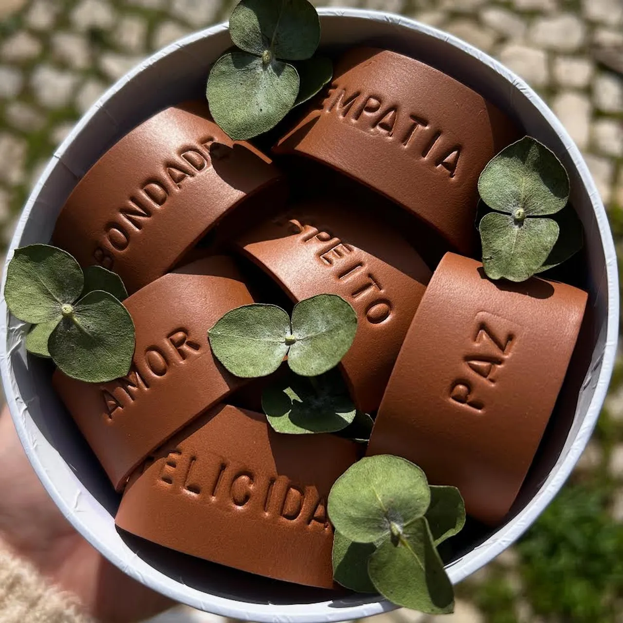 Argolas de guardanapo em couro castanho com palavras gravadas como “Bondade”, “Empatia”, “Amor”, “Respeito”, “Felicidade” e “Paz”, decoradas com pequenas folhas verdes dentro de uma caixa branca.
