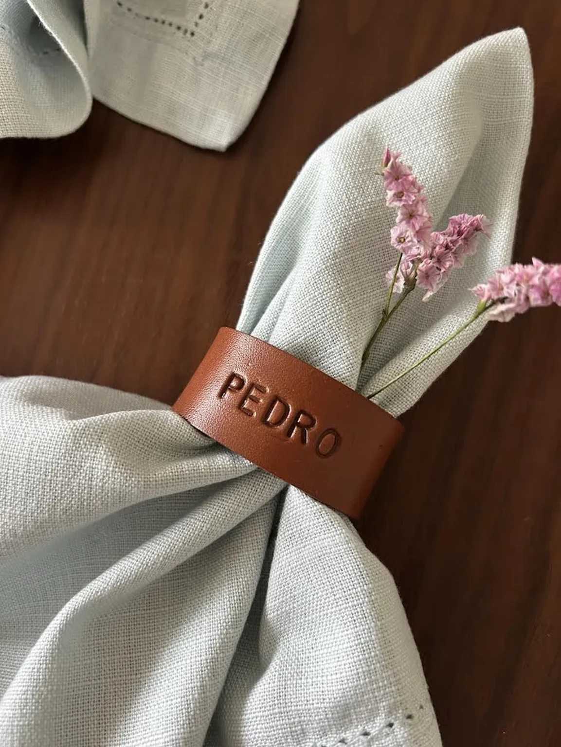 Argola de guardanapo em couro castanho personalizada com o nome “PEDRO”, envolvendo um guardanapo de tecido claro, decorado com pequenas flores rosa.