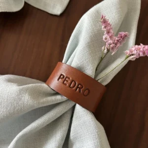 Argola de guardanapo em couro castanho personalizada com o nome “PEDRO”, envolvendo um guardanapo de tecido claro, decorado com pequenas flores rosa.