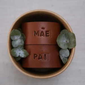Argolas de guardanapo em pele castanha, com as palavras “MÃE” e “PAI”, colocadas dentro de uma pequena taça junto a duas flores secas decorativas.