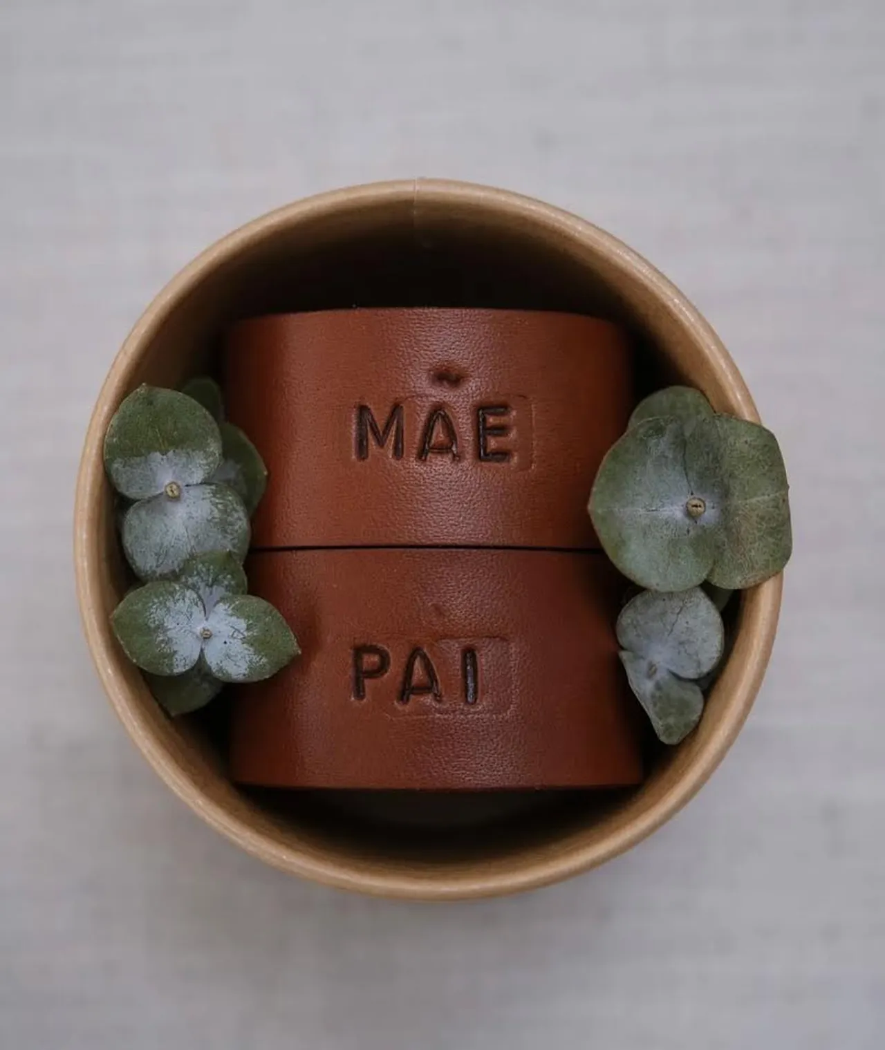 Argolas de guardanapo em pele castanha, com as palavras “MÃE” e “PAI”, colocadas dentro de uma pequena taça junto a duas flores secas decorativas.