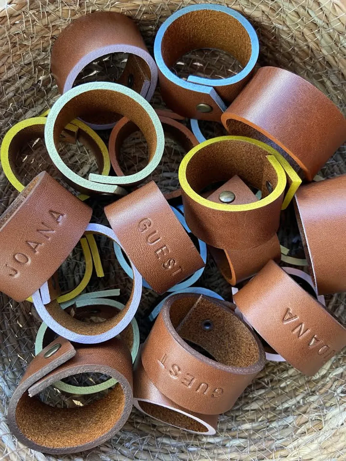 Argolas de guardanapos em couro personalizadas com nomes, em diferentes cores, dentro de um cesto de palha.