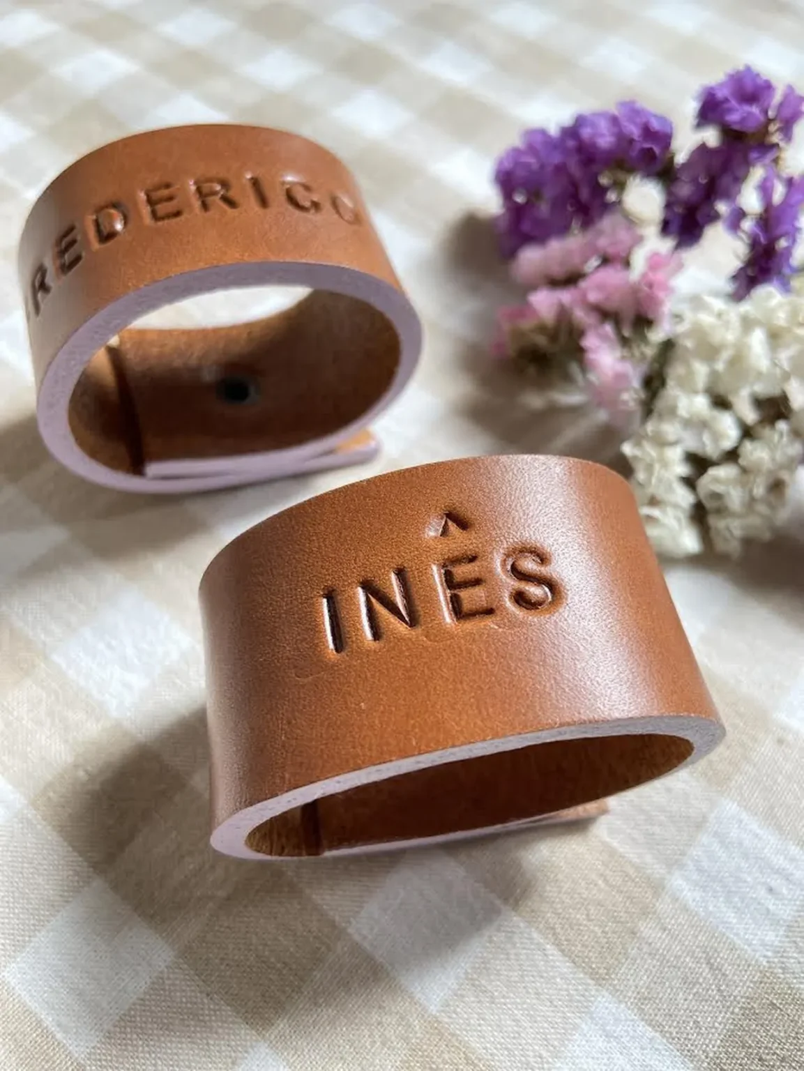 Argolas de guardanapo em couro castanho personalizadas com nomes gravados, apresentadas sobre um fundo neutro com flores decorativas