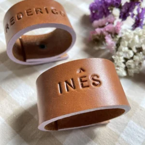 Argolas de guardanapo em couro castanho personalizadas com nomes gravados, apresentadas sobre um fundo neutro com flores decorativas