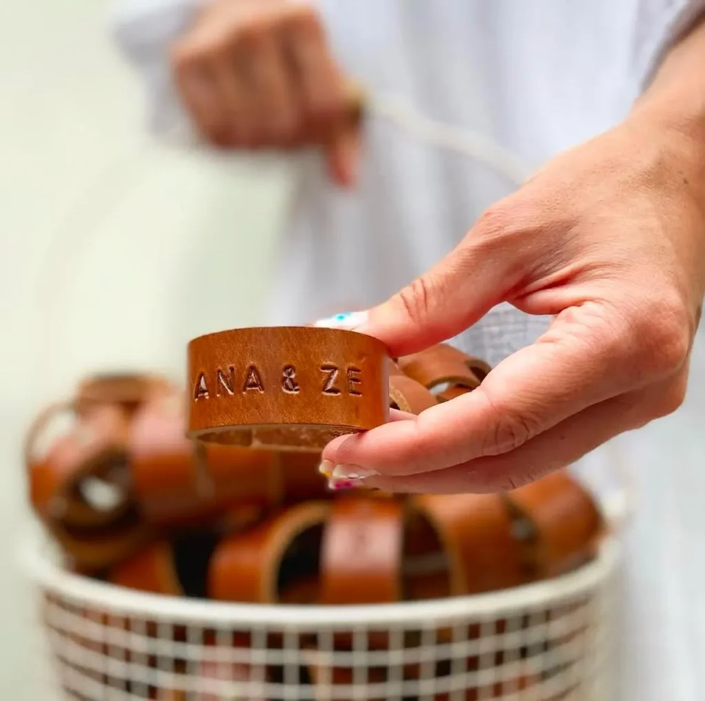 Argola de guardanapo em couro castanho com gravação personalizada “ANA & ZÉ”, segurada por uma mão, com várias argolas dentro de um cesto ao fundo.