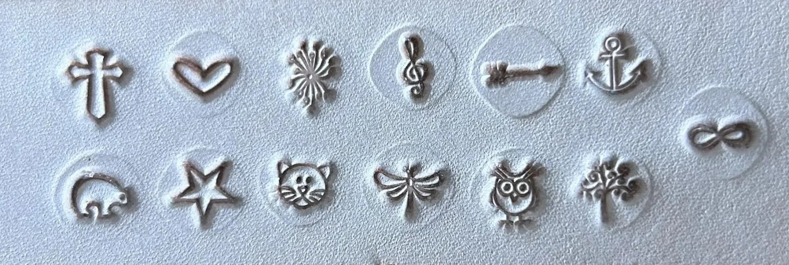 Símbolos em relevo no couro para personalização — cruz, coração, flor, clave de sol, âncora, infinito, estrela, gato, libélula, coruja e árvore.